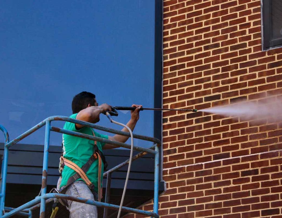 A worker on a lift using a pressure washer to clean the exterior of a building, demonstrating Building Pressure Washing Services to maintain the cleanliness and appearance of commercial structures.