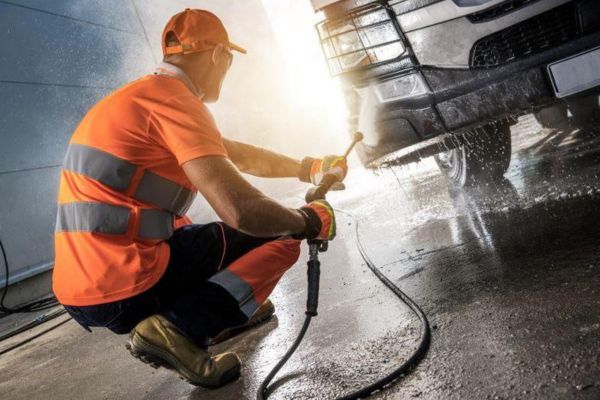 A technician in high-visibility workwear performs commercial pressure washing services, cleaning the front of a vehicle with a high-pressure hose in a professional facility.