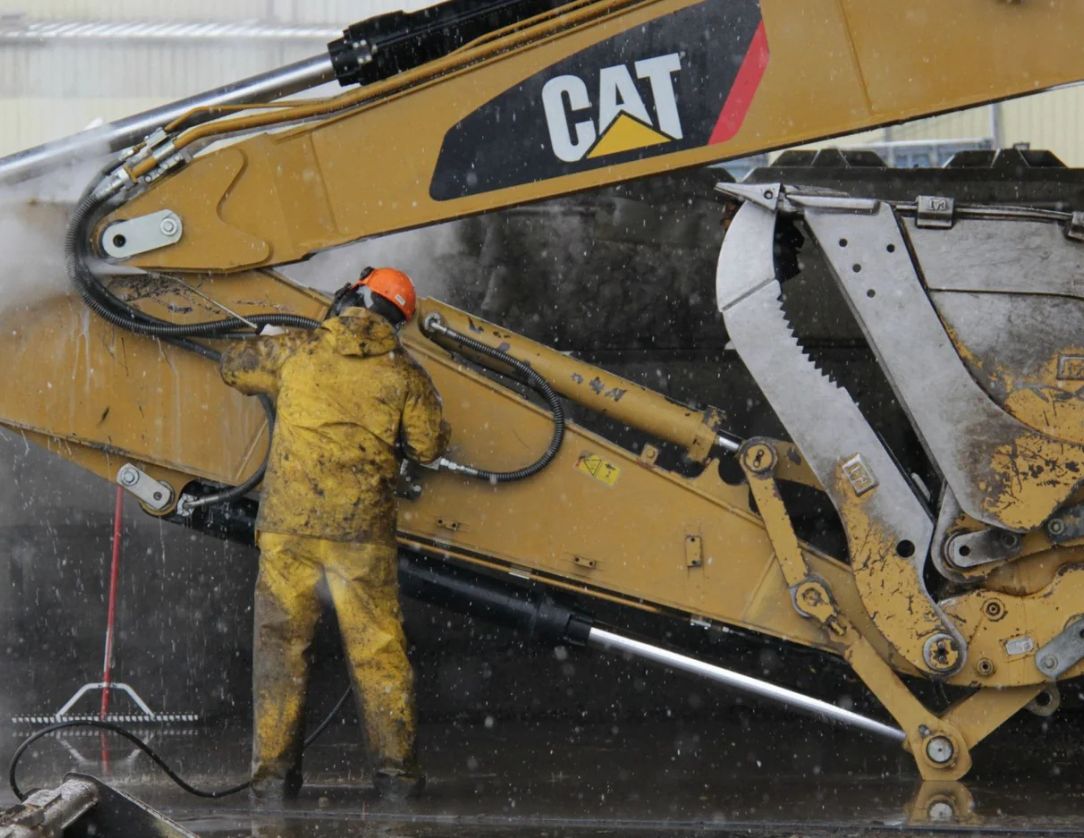 A worker in protective gear pressure washing a CAT excavator, showcasing Excavator Pressure Washing Services to ensure machinery cleanliness and functionality.