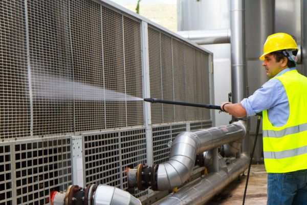 A worker wearing safety gear performs industrial pressure washing services, using high-pressure water to clean metal surfaces and equipment at an industrial facility.