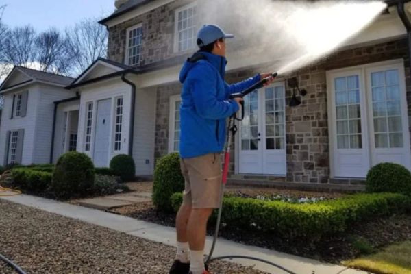 A professional performing residential pressure washing services, spraying high-pressure water to clean the exterior walls of a modern home with stone and siding finishes.