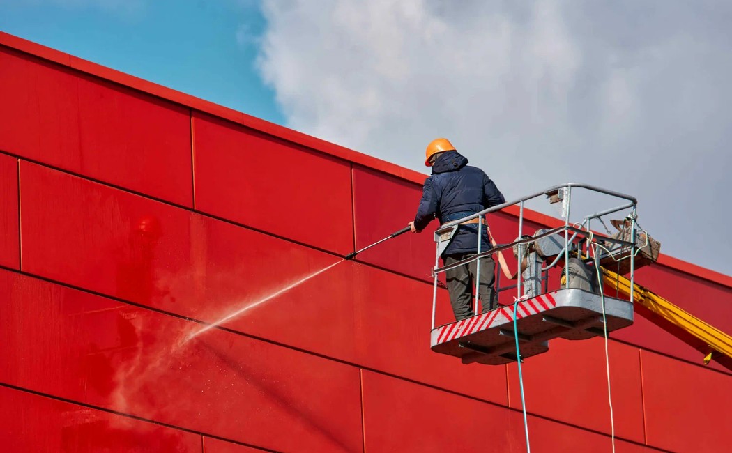 A worker using professional Building Washing Services to clean a large red commercial exterior wall with a pressure washer from an elevated platform.