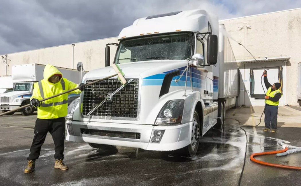 Workers from a Fleet Washing Company clean a large white semi-truck using brushes and pressure washers in an industrial area.
