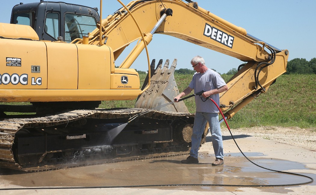 A worker uses a high-pressure washer to clean the tracks of a large excavator outdoors, demonstrating services provided by a Heavy Equipment Washing Company.