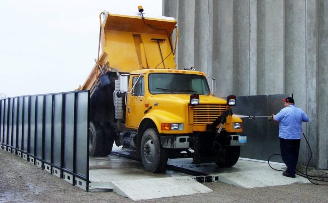 A worker uses a high-pressure washer to clean a large yellow dump truck at an outdoor wash station, demonstrating professional Heavy Equipment Washing Services.