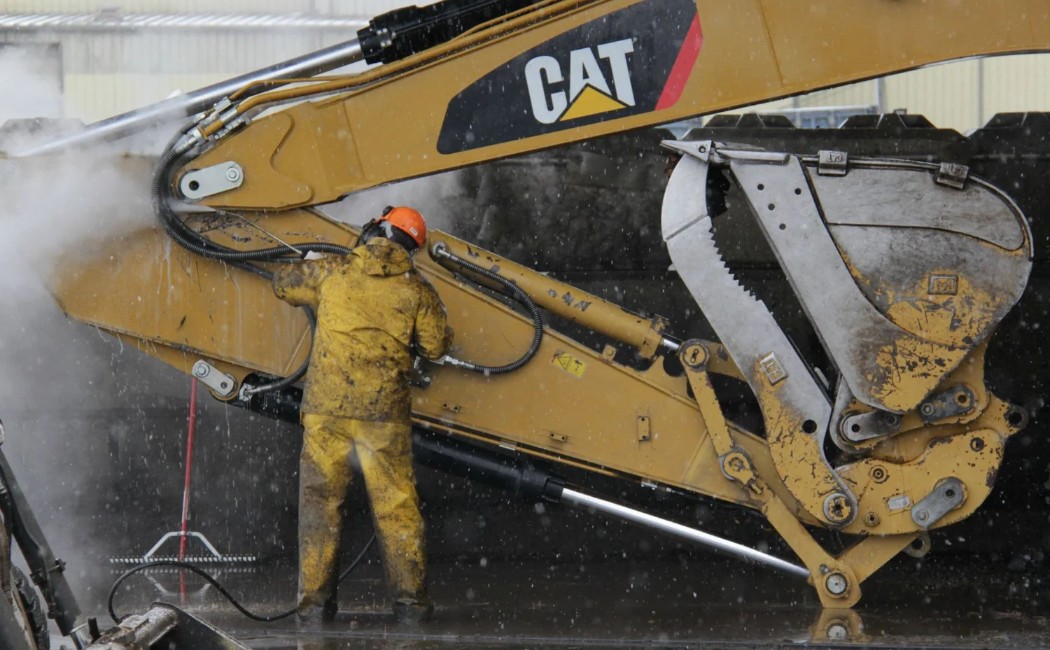 A worker in protective gear is pressure washing a CAT excavator, demonstrating Excavator Washing Services to maintain heavy machinery.