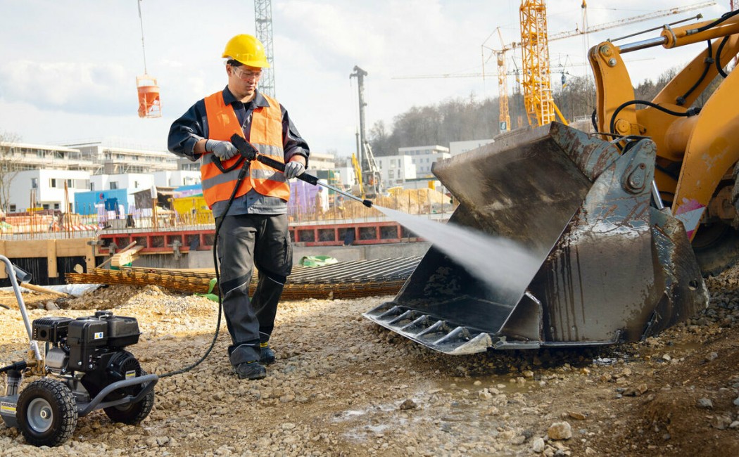 A worker uses a pressure washer to clean an excavator's bucket at a construction site, ensuring proper maintenance of heavy machinery.