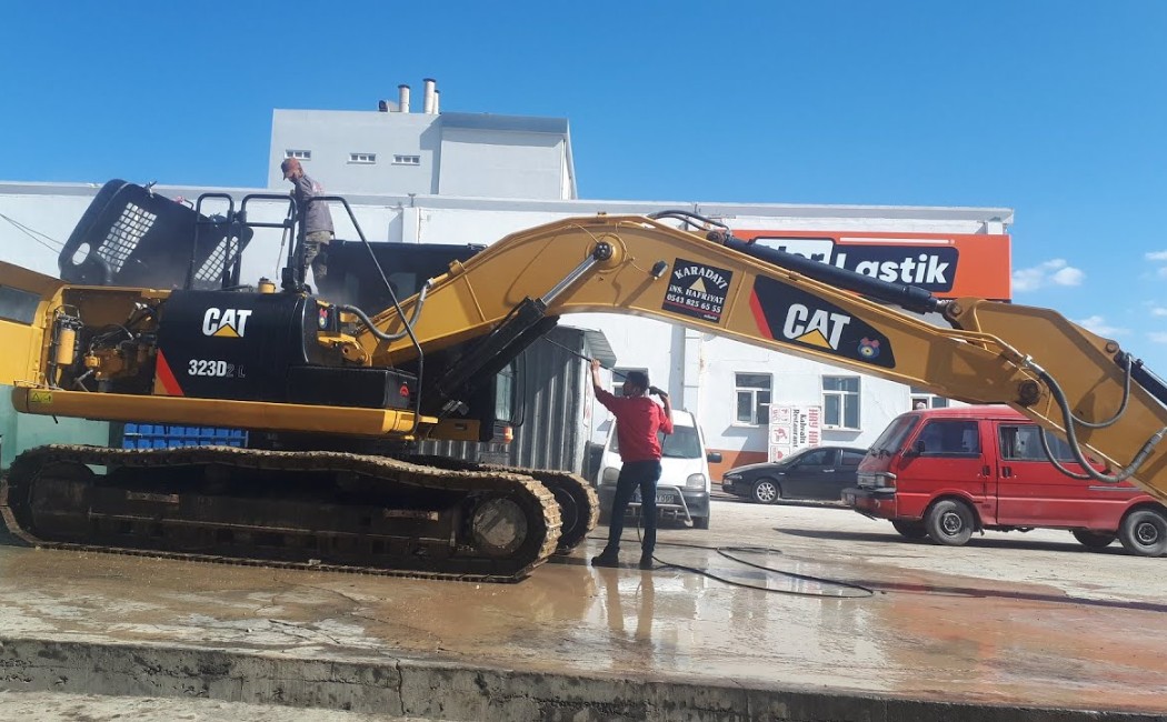 A worker performs professional excavator washing on a CAT 323D2 L at a service area, using high-pressure water to clean the machinery thoroughly.