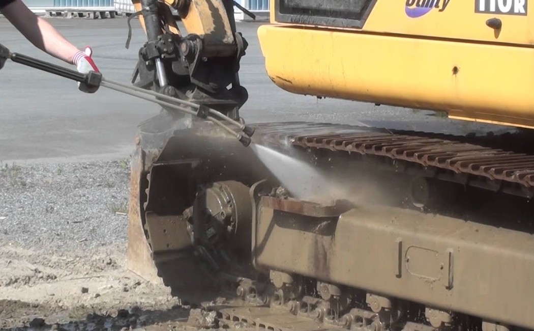 A worker is performing professional pressure washing on an excavator's undercarriage, using high-pressure water to remove dirt and debris.