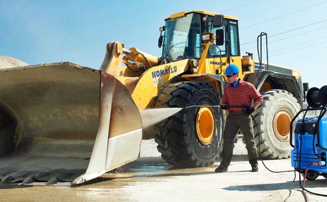 Industrial Heavy Equipment Pressure Washing as a worker uses a high-pressure washer to clean a large front loader at an industrial site, removing dirt from the bucket and tires under clear daylight.