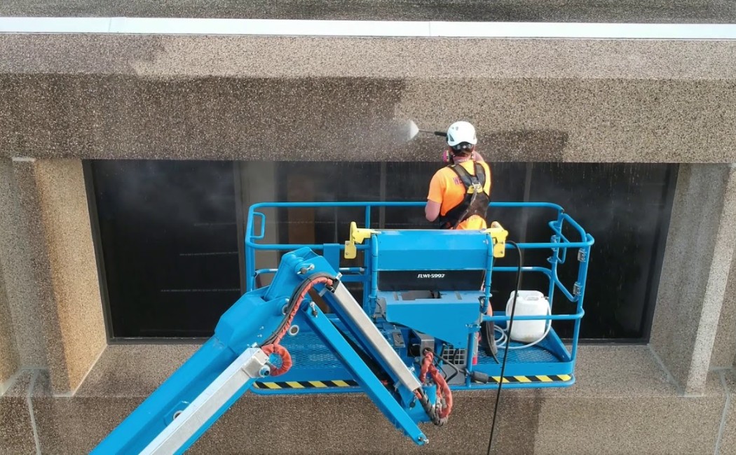 Technician using a lift to pressure wash the exterior façade of a commercial structure, delivering safe and effective Building Washing Services to remove dirt and stains.