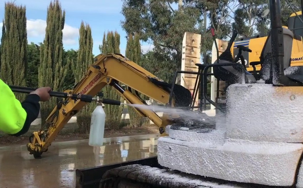 A worker performing Industrial Excavator Pressure Washing to remove heavy dirt and grease from construction machinery using high-pressure water and foam cleaning.