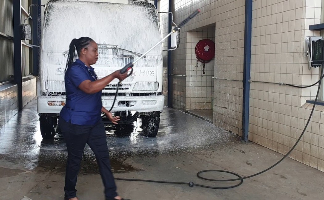 Industrial Fleet Pressure Washing in action as a worker uses a high-pressure hose to clean a truck inside a vehicle wash bay, with foam covering the truck’s front and wet floors below.