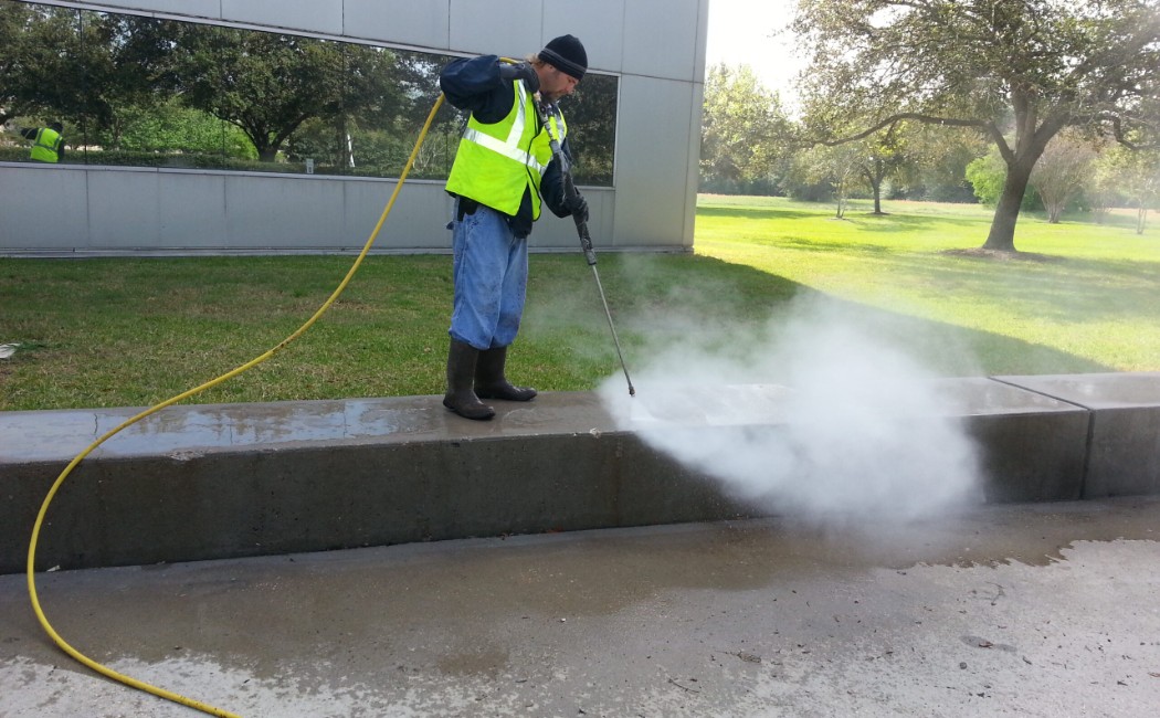 Professional worker pressure washing a concrete curb and walkway outside a commercial building, showcasing Commercial Pressure Washing Services for exterior surface cleaning.